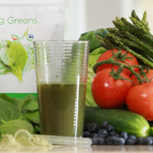 Vaso de Giving Greens preparado, rodeado de verduras verdes frescas con el paquete de Giving Greens visible al fondo