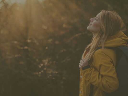Image d'une femme en manteau dans les bois qui ferme les yeux et profite du soleil d'automne